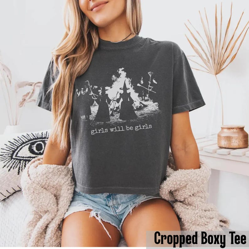 Pu&ograve; includere: Una t-shirt a maniche corte color grigio antracite con una stampa grafica bianca raffigurante sagome di streghe attorno a un fal&ograve;. Il testo "girls will be girls" &egrave; stampato in bianco sotto la grafica. Il capo ha un taglio corto e squadrato.