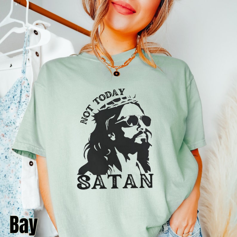 Pu&ograve; includere: Una maglietta verde chiaro con una grafica nera di Ges&ugrave; che indossa occhiali da sole e una corona di spine. Il testo "NOT TODAY SATAN" &egrave; stampato sulla maglietta.