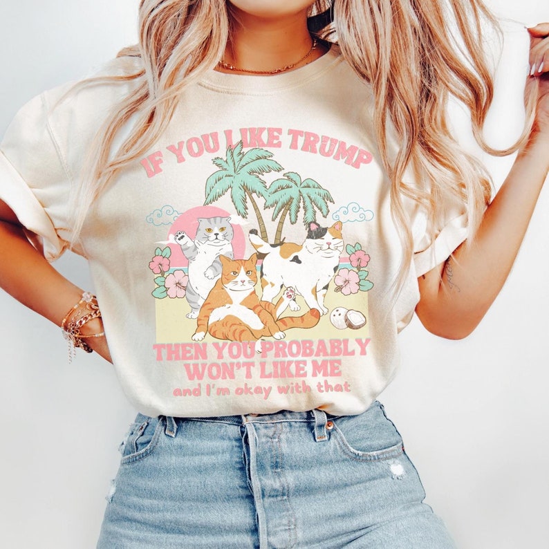 May include: A light beige t-shirt with a graphic print of three cats sitting on a beach with palm trees. The text on the shirt reads "If you like Trump then you probably won't like me and I'm okay with that."