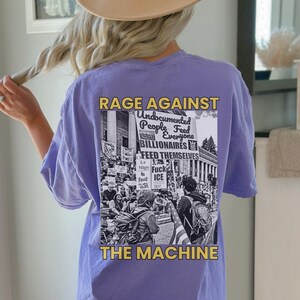 May include: A light purple t-shirt with the text "RAGE AGAINST THE MACHINE" in gold and white. The shirt features a black and white graphic of a protest with the words "Undocumented People Feed Everyone BILLIONAIRES FEED THEMSELVES".
