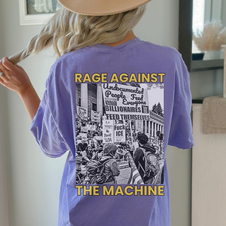 May include: A light purple t-shirt with the text "RAGE AGAINST THE MACHINE" in gold and white. The shirt features a black and white graphic of a protest with the words "Undocumented People Feed Everyone BILLIONAIRES FEED THEMSELVES".