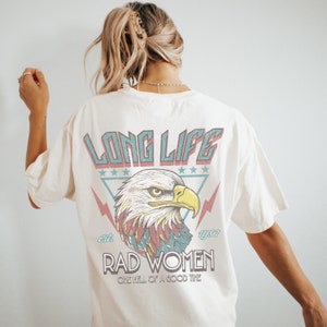 Op de afbeelding: Een wit T-shirt met een vintage-stijl grafische afbeelding van een adelaar met de tekst "Long Life Est. 1977 Rad Women One Hell of a Good Time" gedrukt op de achterkant.