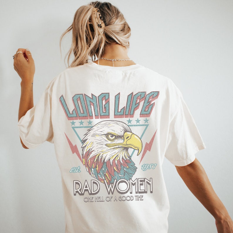 Op de afbeelding: Een wit T-shirt met een vintage-stijl grafische afbeelding van een adelaar met de tekst "Long Life Est. 1977 Rad Women One Hell of a Good Time" gedrukt op de achterkant.