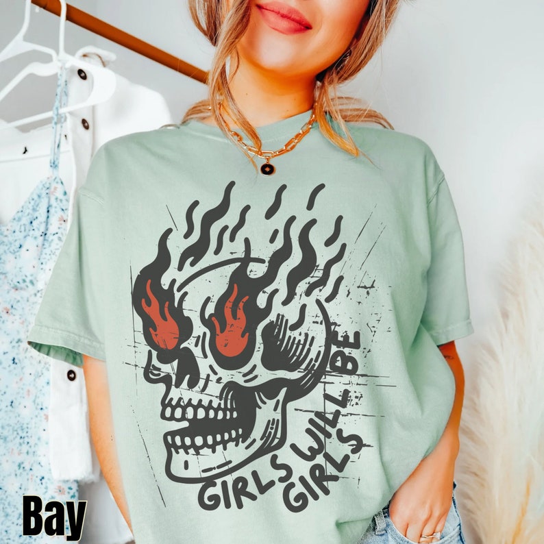May include: A light green t-shirt featuring a black and white skull graphic. The skull has flames for eyes and the text "Girls Will Be Girls" is printed below the skull.