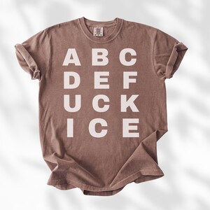 May include: A brown t-shirt with the word "ABC DEF UCK ICE" printed in white block letters. The shirt has a crew neck and short sleeves, and is made of a soft, comfortable material.