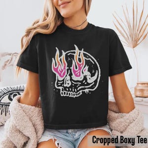 May include: Black cropped boxy tee featuring a white skull graphic with pink and orange flames. The skull design is centered on the front of the shirt. The text "Cropped Boxy Tee" is at the bottom of the image.