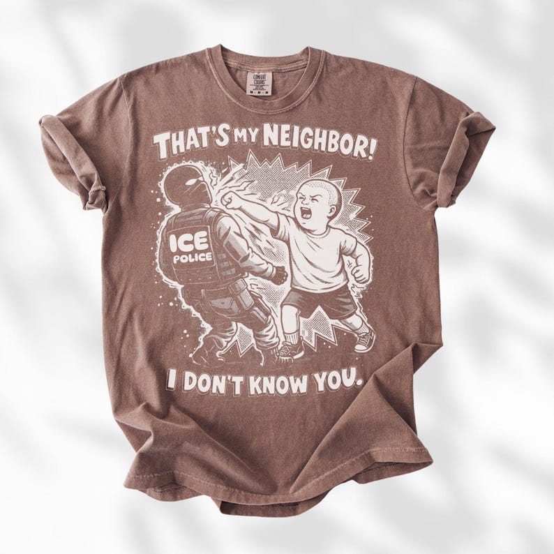 K&ouml;nnte beinhalten: Braunes T-Shirt mit einer Grafik eines Kindes, das einen ICE-Polizisten schl&auml;gt. Der Text auf dem Shirt lautet: "THAT'S MY NEIGHBOR! I DON'T KNOW YOU."