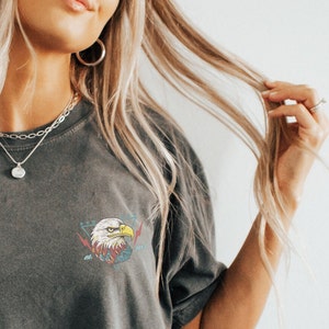 Op de afbeelding: Een grijs T-shirt met een afbeelding van een adelaarskop in een driehoek met bliksemschichten en de tekst "2021" op de voorkant.