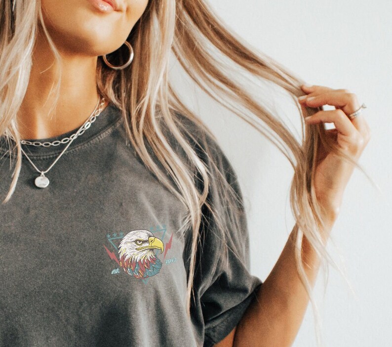 Op de afbeelding: Een grijs T-shirt met een afbeelding van een adelaarskop in een driehoek met bliksemschichten en de tekst "2021" op de voorkant.