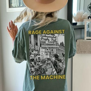 May include: Sage green t-shirt with a graphic on the back that reads "RAGE AGAINST THE MACHINE." The graphic includes text and a black and white image of a protest. The shirt is a casual, oversized fit.