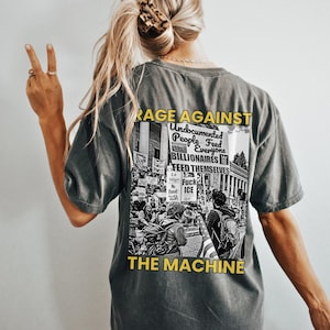 May include: Dark gray t-shirt with a graphic on the back that reads "RAGE AGAINST THE MACHINE." The graphic includes text about undocumented people, billionaires, and ICE. The shirt has a vintage, worn look.