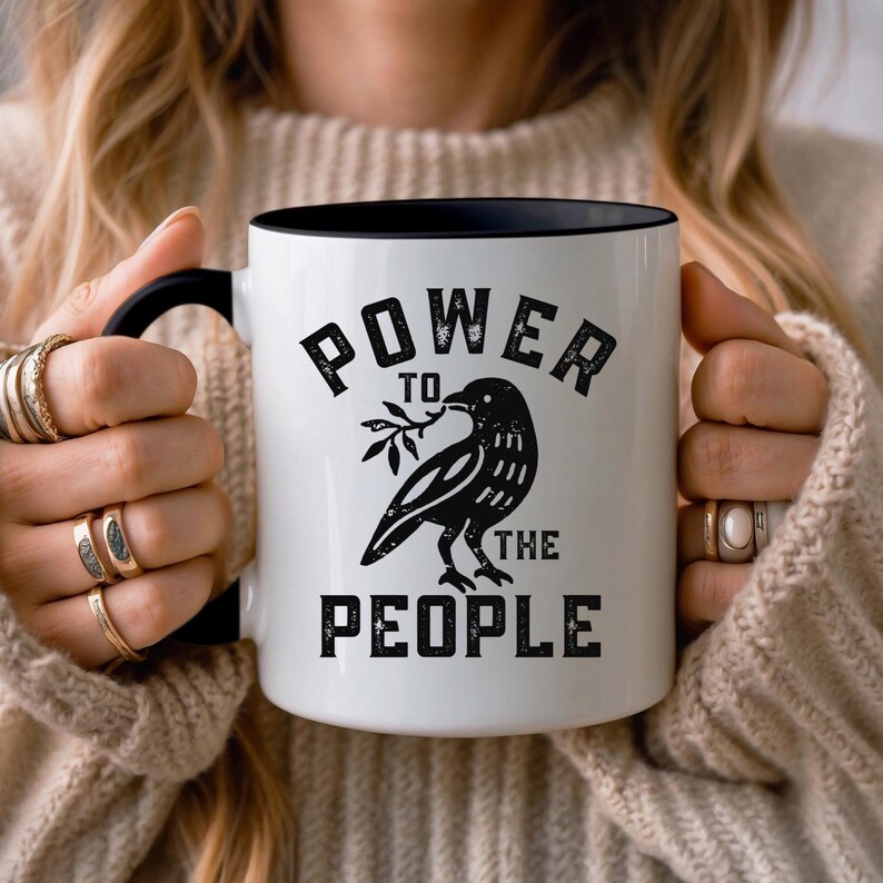 May include: White ceramic mug with a black interior and handle. The mug features a black illustration of a raven holding a sprig, with the words "POWER TO THE PEOPLE" in black lettering. A perfect gift for anyone.