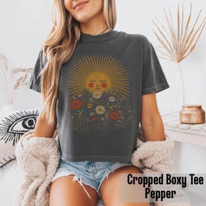 Puede incluir: Una camiseta corta y cuadrada de color gris oscuro con un gr&aacute;fico de sol y flores. El sol tiene una cara y rayos amarillos. Las flores son rojas, blancas y moradas. El texto "Cropped Boxy Tee Pepper" est&aacute; en la parte inferior.
