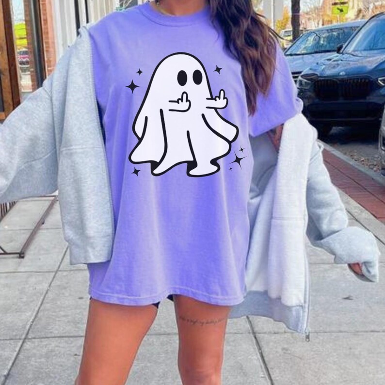 May include: A lavender t-shirt featuring a white ghost graphic giving the middle finger. The ghost has black eyes and is surrounded by four black starbursts. The shirt is worn with a gray zip-up hoodie.