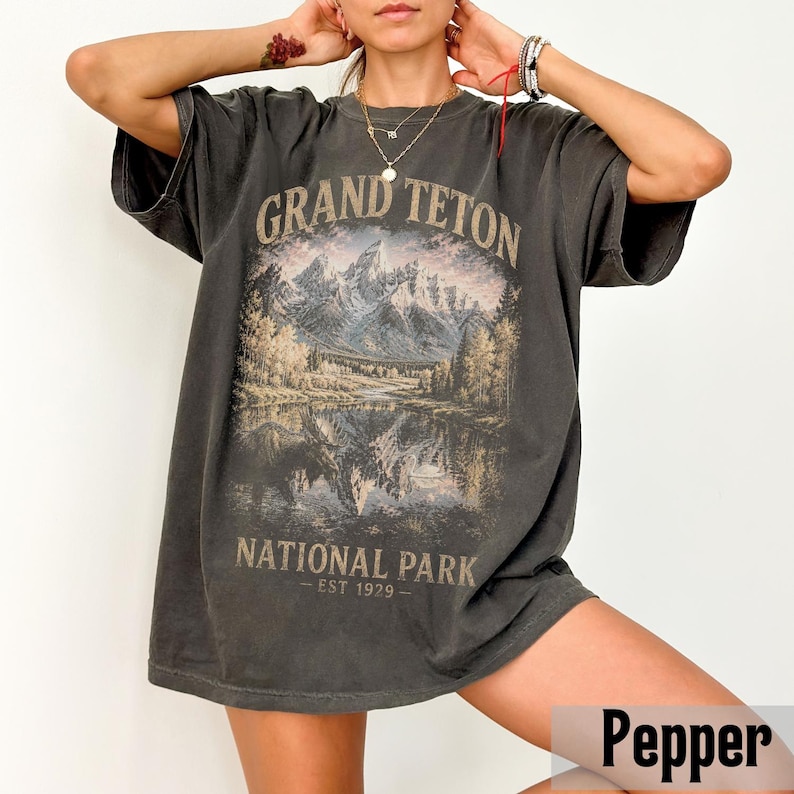 May include: A dark gray t-shirt featuring a scenic mountain and lake graphic with the text "GRAND TETON NATIONAL PARK - EST 1929". The shirt has short sleeves and a relaxed fit, perfect for casual wear.