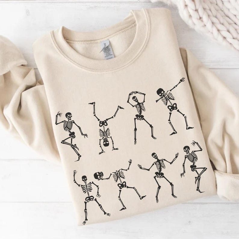K&ouml;nnte beinhalten: Beigefarbenes Sweatshirt mit einem schwarzen Aufdruck tanzender Skelette. Die Skelette befinden sich in verschiedenen Posen, mit ausgestreckten Armen und Beinen. Das Sweatshirt ist aus weichem Material, ideal f&uuml;r Halloween.