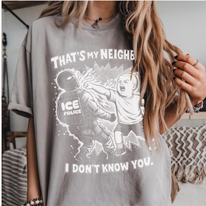 K&ouml;nnte beinhalten: Graues T-Shirt mit einer Grafik, die ein Kind zeigt, das einen ICE-Polizisten schl&auml;gt. Der Text auf dem Shirt lautet: "THAT'S MY NEIGHBOR, I DON'T KNOW YOU."