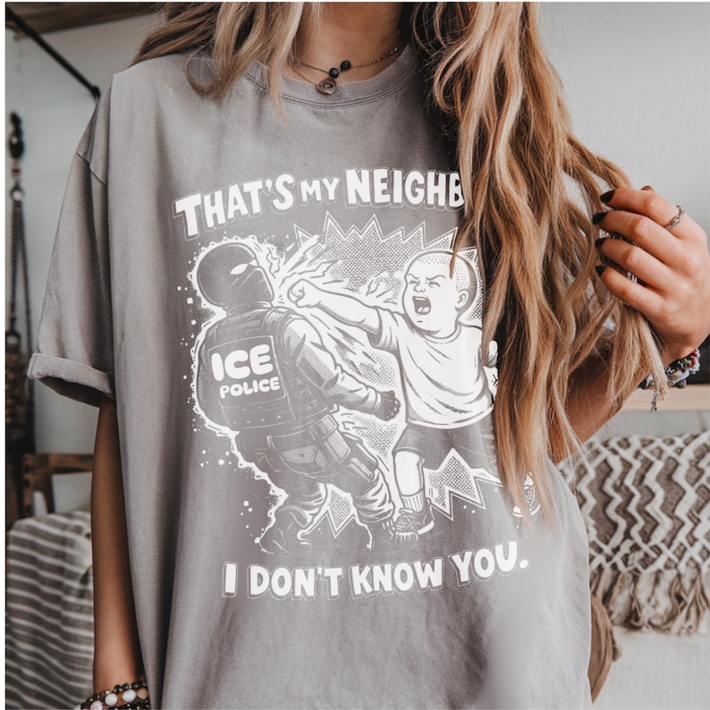 K&ouml;nnte beinhalten: Graues T-Shirt mit einer Grafik, die ein Kind zeigt, das einen ICE-Polizisten schl&auml;gt. Der Text auf dem Shirt lautet: "THAT'S MY NEIGHBOR, I DON'T KNOW YOU."