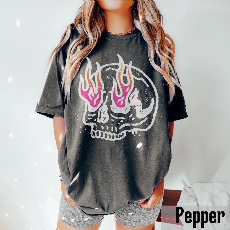 May include: Dark gray t-shirt featuring a white skull graphic with pink and orange flames. The graphic tee has a vintage, distressed look. The shirt is a relaxed fit and has short sleeves. The word "Pepper" is in the bottom right corner.