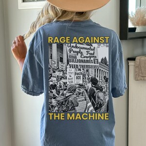 May include: Blue short-sleeved t-shirt with a graphic on the back. The graphic features the text "RAGE AGAINST THE MACHINE" in yellow, with a black and white image of a protest. The image includes the text "Undocumented People Feed Everyone Billionaires".