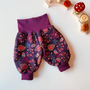 Op de afbeelding: Donkerpaarse babybroek met een hoge tailleband en manchetten. De broek heeft een kleurrijk bloemenpatroon met rode, oranje en blauwe accenten op een donkerpaarse achtergrond. Gemaakt van een zacht, comfortabel materiaal.