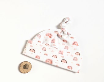 May include: A white baby hat with a knotted top. The hat is covered in a repeating pattern of pink and brown rainbows with small hearts and wavy lines.