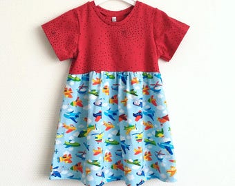May include: A red and blue toddler dress with a playful airplane pattern. The top is solid red with small black dots, and the skirt features colorful airplanes on a light blue background. The text "Size 2T / Maat 92" is visible.