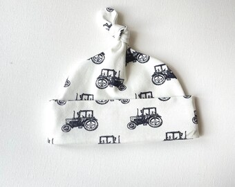 May include: White baby hat with a navy blue tractor pattern. The hat is made of soft fabric and features a knotted top. The tractor design is repeated across the hat's surface, creating a charming and playful look. A perfect accessory for a newborn.