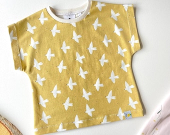 May include: Mustard yellow short-sleeved top featuring a repeating white bird design. The shirt has a cream-coloured neckline and the text "Size 12-18M / Maat 80-86" at the top. Made from a soft fabric.
