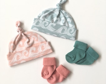 May include: Two sets of baby socks and two baby hats with a wave pattern. One set of socks is teal and the other is pink. One hat is light blue and the other is pink.