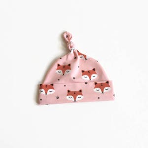 May include: A pink baby hat with a knot on top. The hat has a pattern of orange fox faces with closed eyes and black dots.