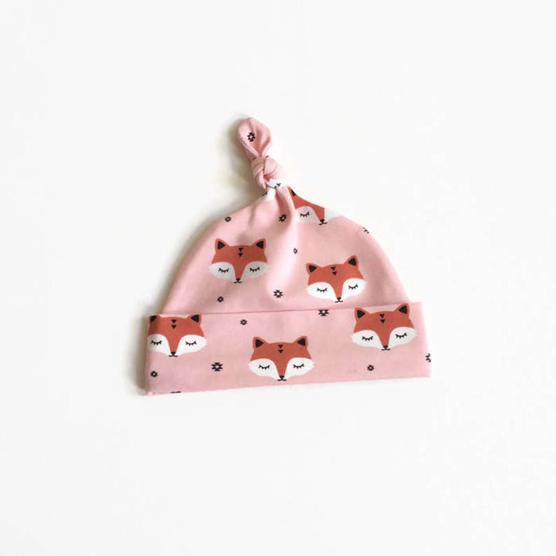 May include: A pink baby hat with a knot on top. The hat has a pattern of orange fox faces with closed eyes and black dots.