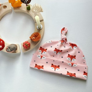 May include: A pink baby hat with a knotted top and a repeating pattern of sleeping foxes. The foxes are orange with black eyes and noses.