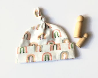 May include: A white baby hat with a tie knot on top. The hat is covered in a pattern of watercolor rainbows in shades of pink, orange, yellow, green, and purple.