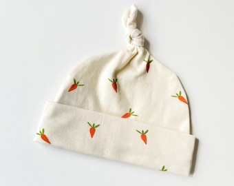 May include: A cream-colored baby hat with a knotted top and a pattern of orange carrots.