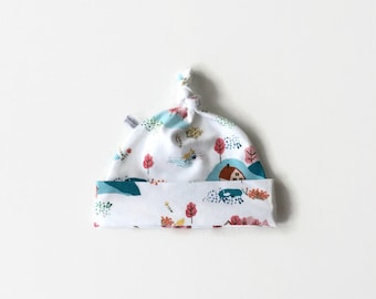 May include: White baby hat with a knotted top. The hat has a colorful pattern of houses, trees, and birds on a white background.