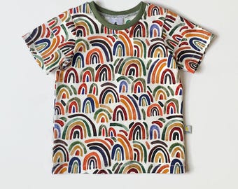 May include: White short-sleeved t-shirt with a repeating rainbow pattern in various colours. The shirt has a green neckline and sleeve trim. The text "Size 4T / Maat 104" is printed at the bottom of the shirt.