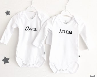 May include: Two white baby onesies with long sleeves, each bearing the name "Anna" in black script. The onesies hang on white hangers against a white background, with gray star decorations.