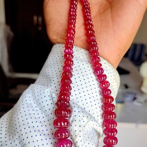 May include: A strand of deep red ruby beads, each bead is carved with a flower-like design. The beads are strung on a gold thread and are held in a hand.