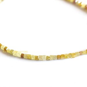 May include: A delicate bracelet composed of small, square-cut beads in varying shades of yellow, ranging from pale lemon to deep amber. The beads are strung together in a single strand, featuring a gold-toned metal clasp at the end of the chain.