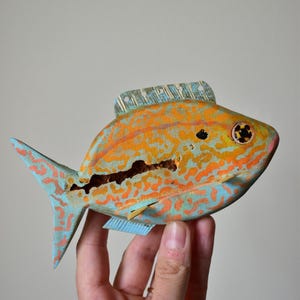 Könnte beinhalten: Eine handgeschnitzte Holzfischskulptur mit einem lebendigen orangefarbenen und hellblauen gesprenkelten Muster. Der Fisch hat eine markante Rückenflosse, ein detailliertes Auge und einen sichtbaren Riss quer über den Körper.