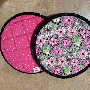 May include: Two round quilted pot holders. One is bright pink with white polka dots and the other has a floral pattern in pink, gray, and black on a gray background. Both have a black border with white dots.