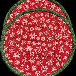 May include: Two round, quilted pot holders. The pot holders have a red background with white snowflakes. The edges are trimmed with a green fabric. The pot holders are designed for use in the kitchen.