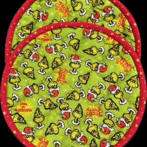 May include: Two round, quilted pot holders with a green background featuring a repeating pattern of a yellow character wearing a Santa hat. The pot holders have a red border with white stars and the text "Feelin' Grinchy" and "Naughty or Nice".
