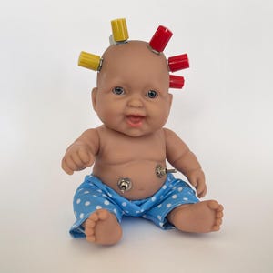 Pode incluir: Uma boneca de bebê com um maiô azul e branco de bolinhas e botões vermelhos e amarelos na cabeça.