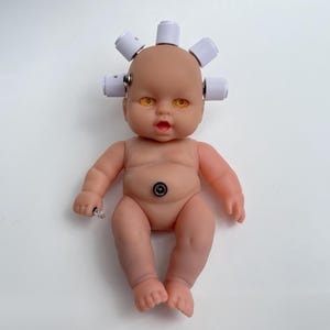 May include: A nude-coloured baby doll with white cylindrical attachments on its head. The doll has yellow eyes and a black and silver button on its stomach. It is holding a small, clear object in its left hand.