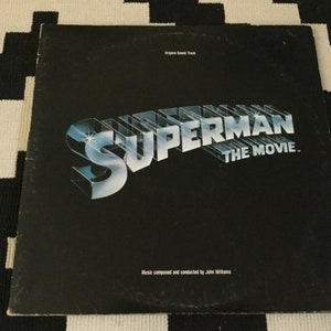 May include: A vintage vinyl record album cover for "Superman: The Movie." The cover is black with the title in a 3D chrome-style font. The text "Original Soundtrack" and the composer's name are also visible.