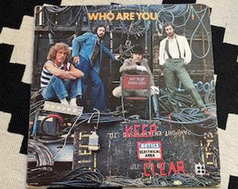 Puede incluir: Portada del álbum "Who Are You" de The Who. La imagen muestra a los miembros de la banda posando frente a un fondo de equipos eléctricos y cables. El título del álbum está en la parte superior y las palabras "KEEP CLEAR" son visibles.