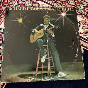 May include: A vinyl record cover titled Richard Pryor's Greatest Hits featuring a photograph of a person sitting on a wooden stool, playing an acoustic guitar in front of a microphone. The background is black with two bright starburst light effects.