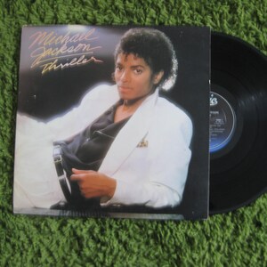 May include: A vinyl record album cover featuring Michael Jackson. The cover art shows Michael Jackson in a white jacket and black shirt, with the album title "Thriller" in gold lettering.